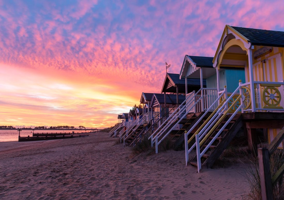 A row of pastel beach huts along Wells-next-the-Sea beach beneath a pink and orange dawn sky