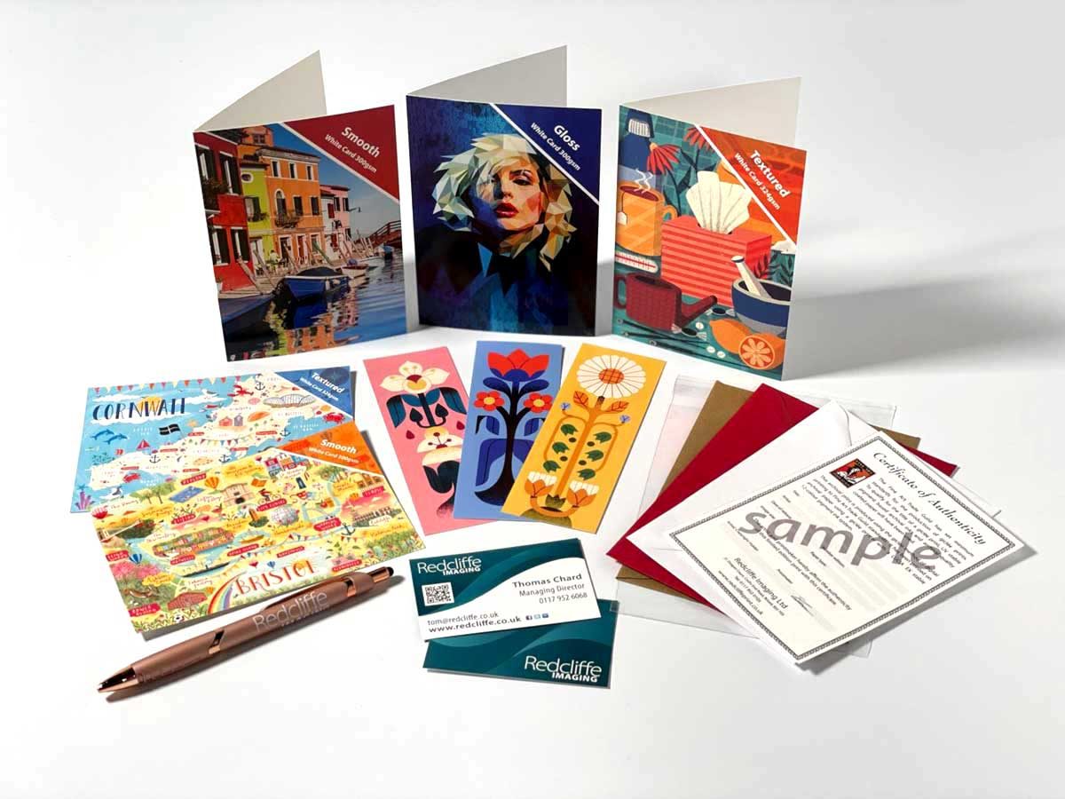 A selection of Redcliffe greeting card products — three card finishes (Smooth, Gloss, Textured), bookmarks, postcards, envelopes, certificate of authenticity, and a Redcliffe pen.